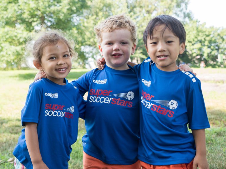 Three young, smiling children in blue soccer uniforms standing side-by-side on a field.