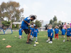 An adult coach speaks to a team of youth soccer players on an outdoor field on dribbling a soccer ball.